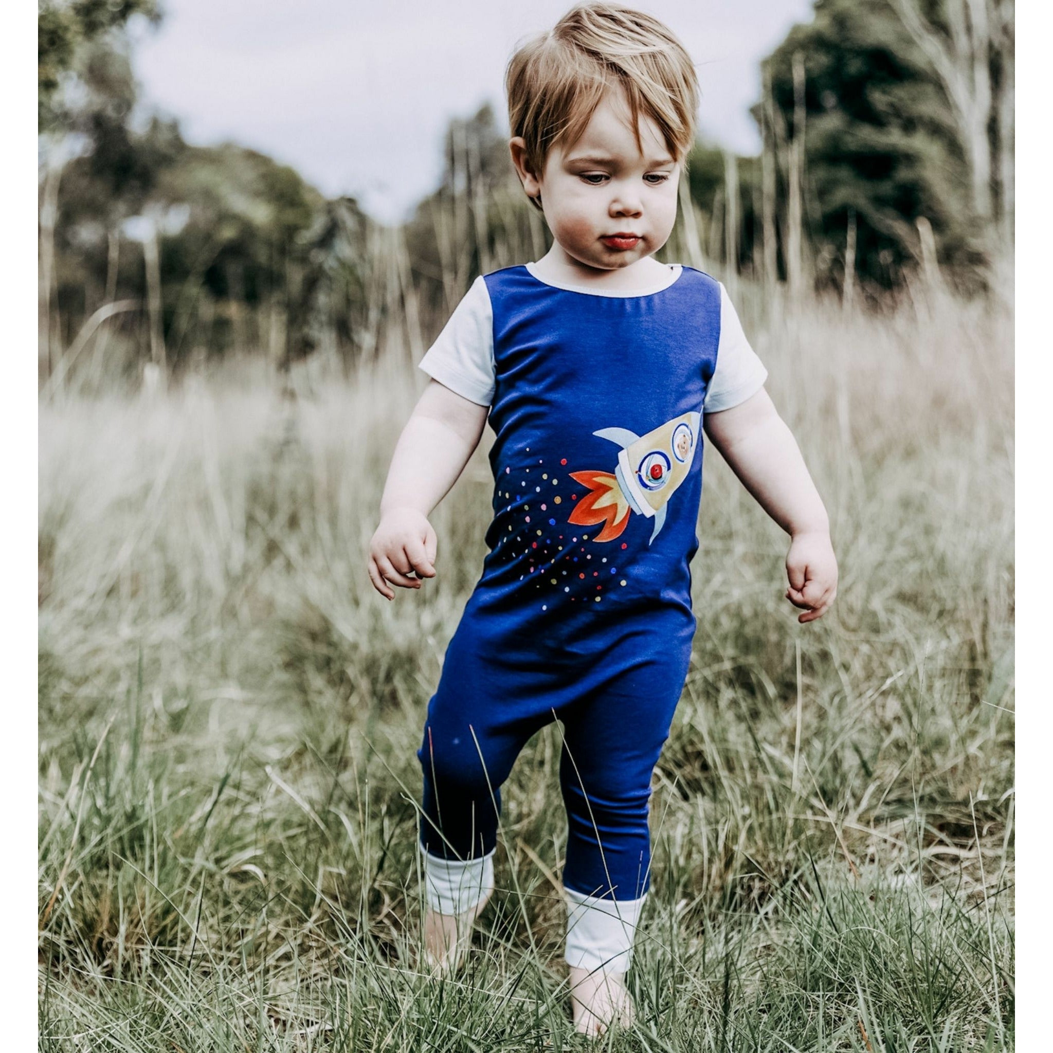 little boy outdoors in grass wearing navy blue romper with rocket illustration